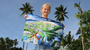 Ein junges Mädchen hält eine farbenfrohe Karte mit dem Titel "Strom in meiner Welt" in der Hand, auf der verschiedene Aktivitäten und Strukturen im Zusammenhang mit Wasser dargestellt sind, vor dem Hintergrund von Palmen und einem klaren blauen Himmel.